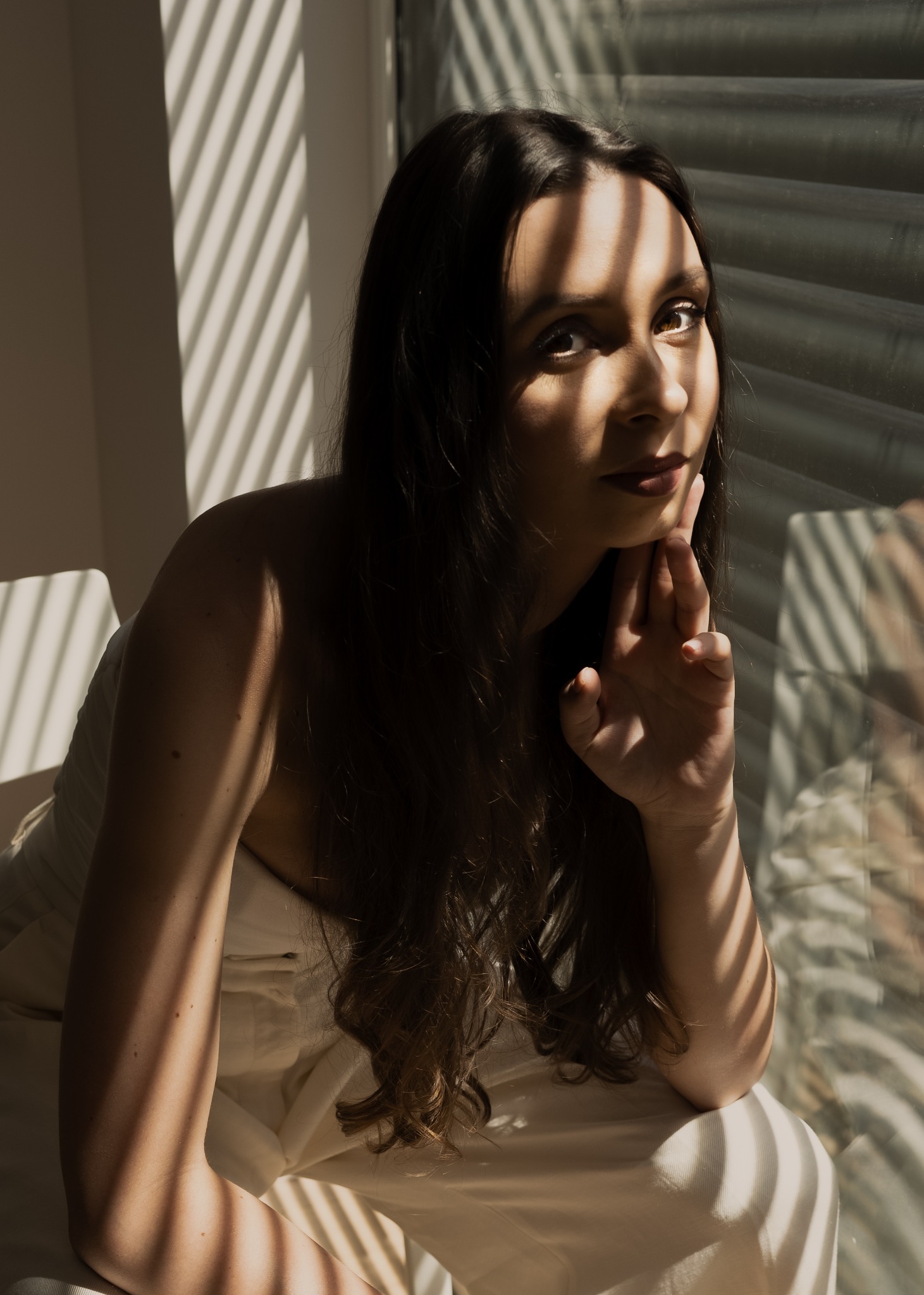 Portret kobiety z długimi ciemnymi włosami, siedzącej w fotelu, oświetlonej przez promienie słoneczne padające przez żaluzje, z ręką podpartą pod brodą i delikatnym uśmiechem.