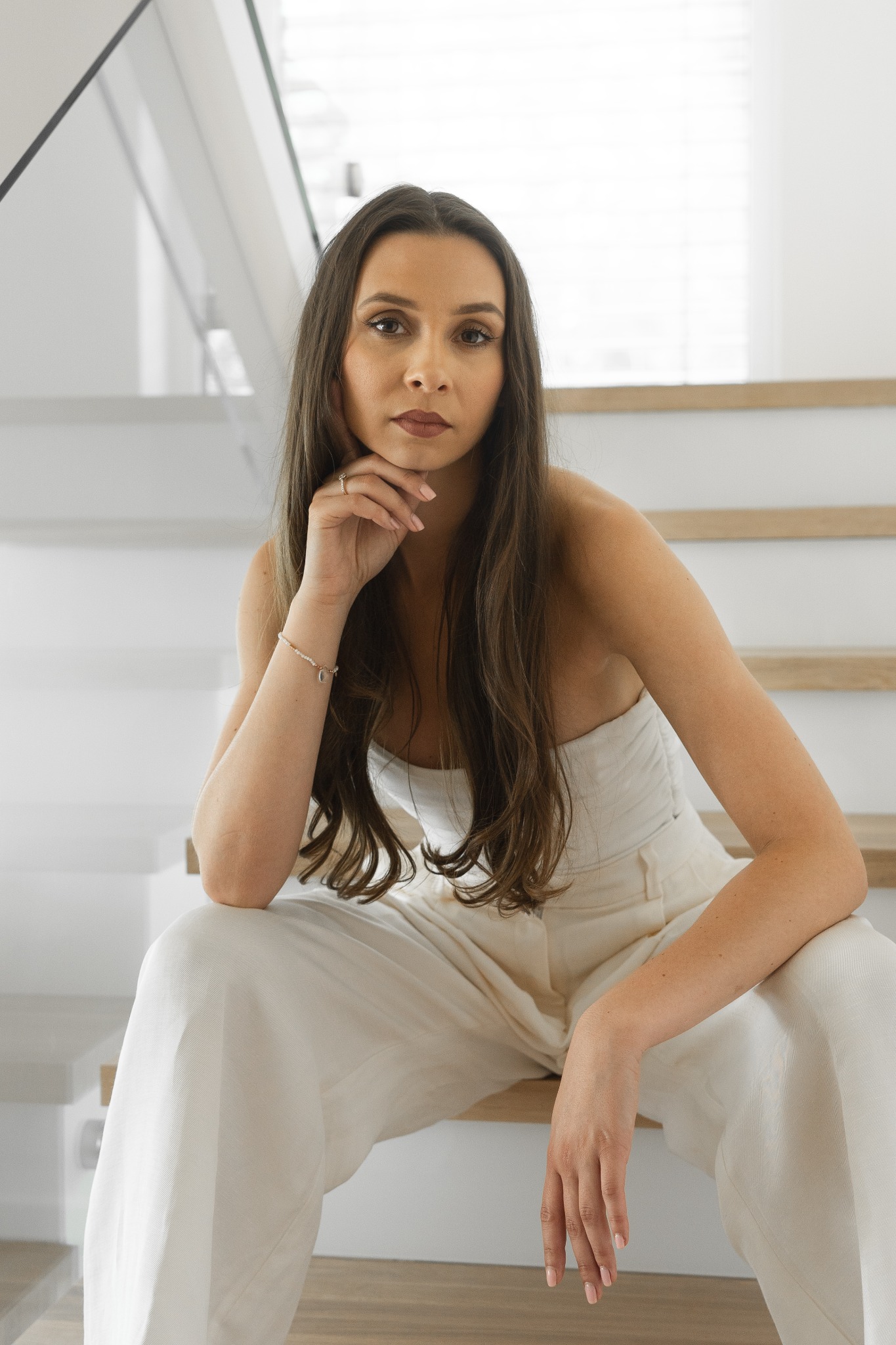 Portret kobiety w białym stroju siedzącej na schodach, z ręką podpartą pod brodą, w minimalistycznym wnętrzu.