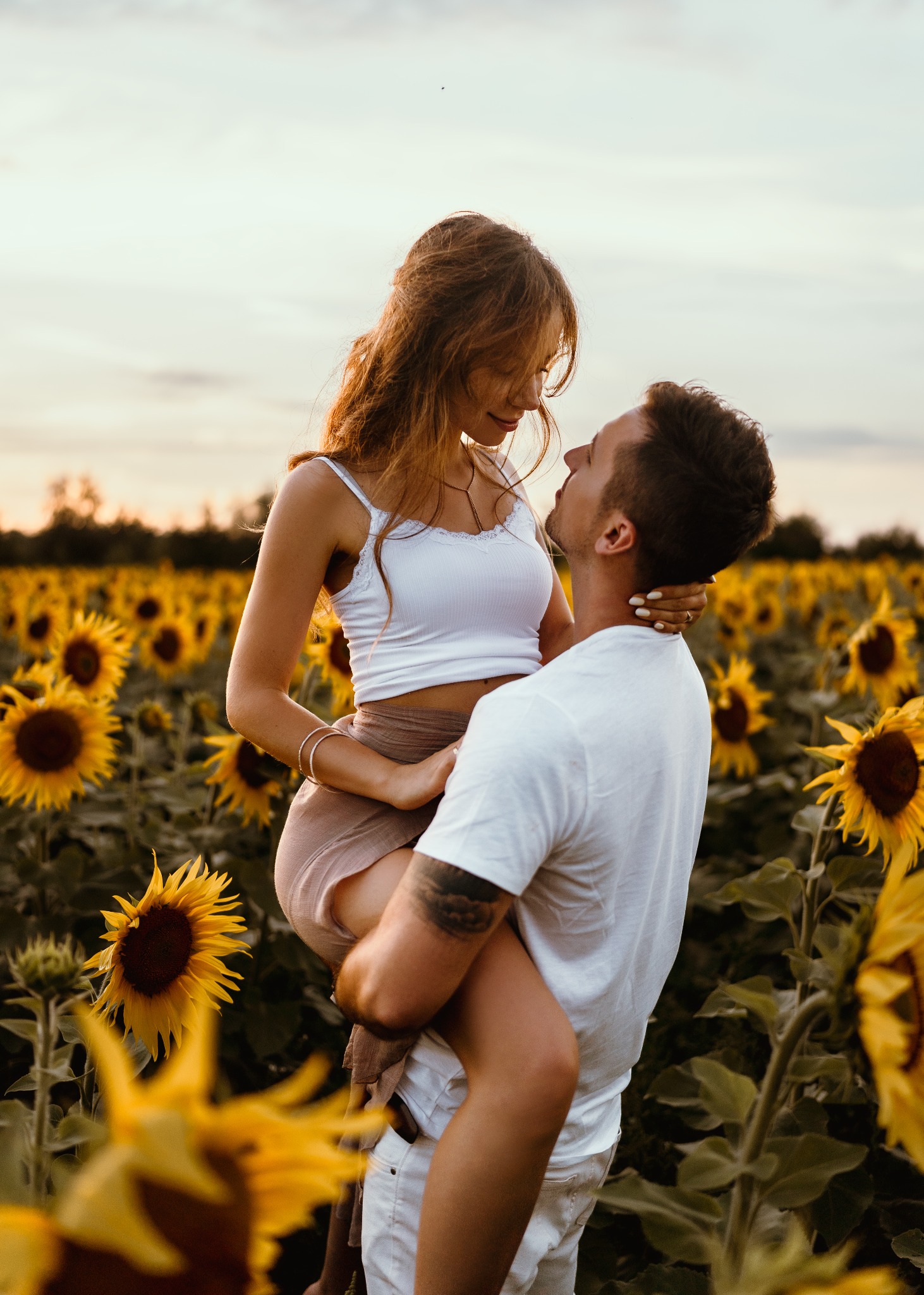 Mężczyzna z tatuażem trzymający kobietę na rękach na tle pola słoneczników, oboje patrzą sobie w oczy.