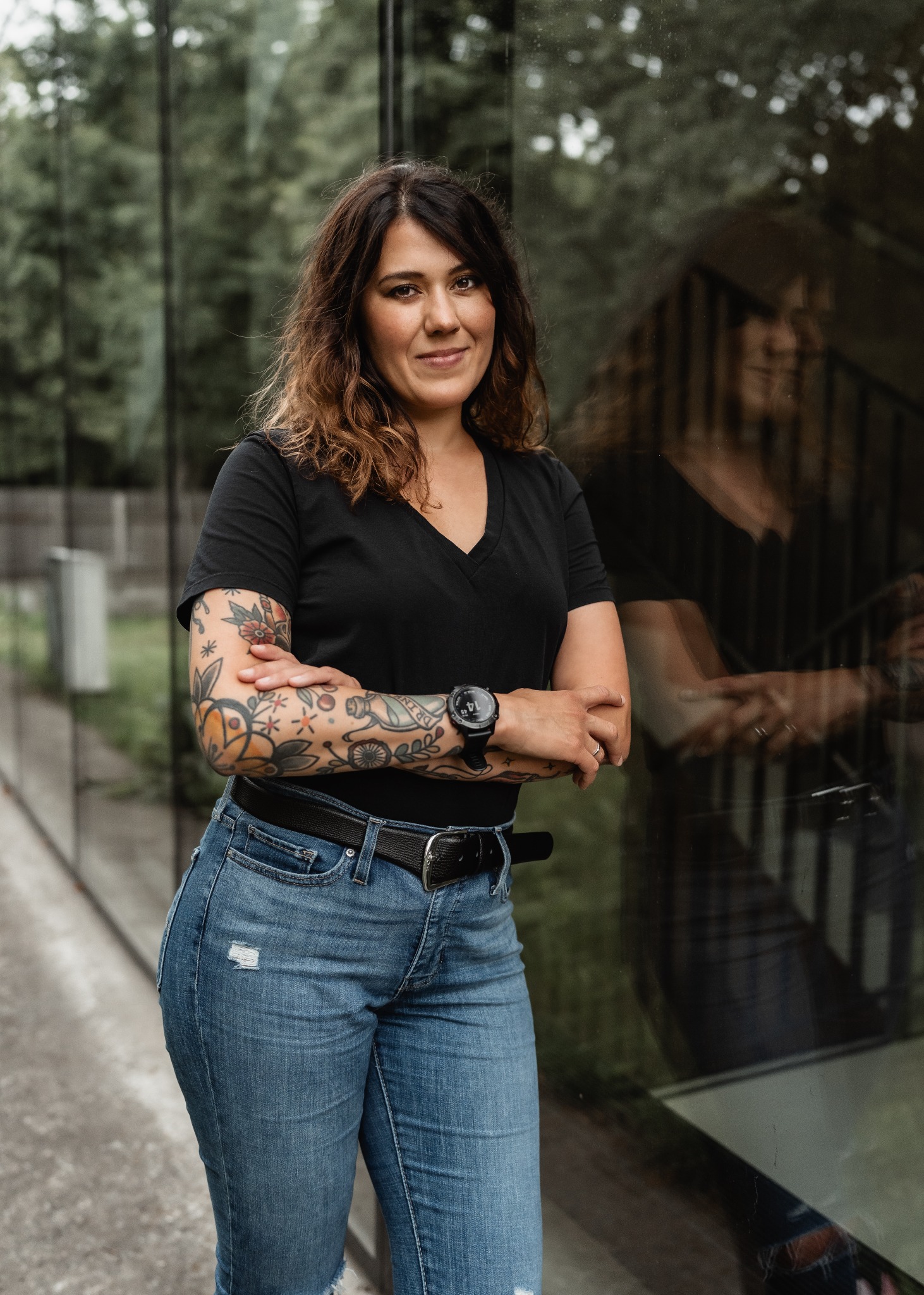 Kobieta z tatuażami na ramionach, ubrana w czarną koszulkę i jeansy, stoi na tle szklanej ściany z odbiciem drzew.