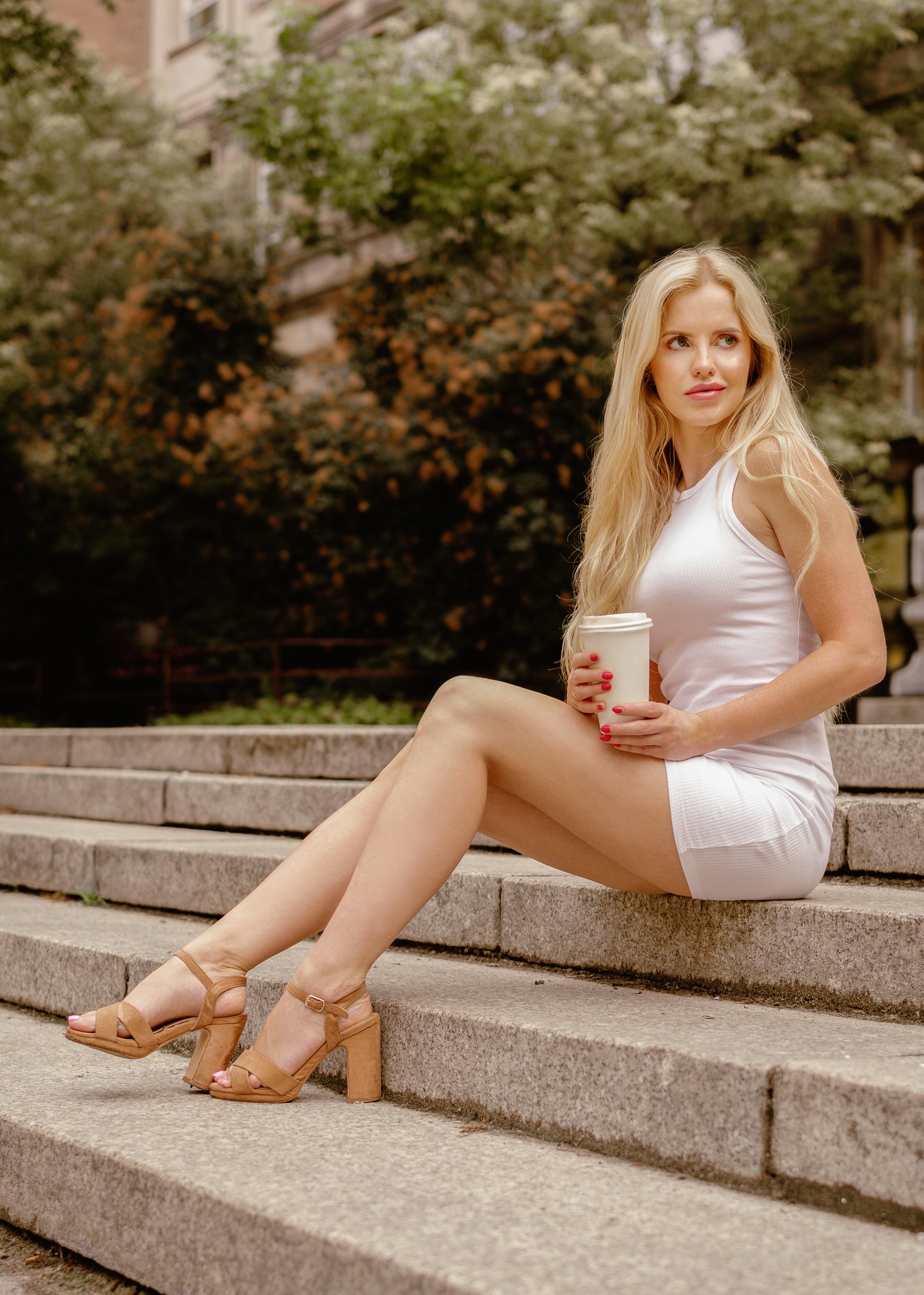 Blondwłosa kobieta w białej sukience siedzi na kamiennych schodach z kawą na wynos, spoglądając w bok, z budynkiem i zielenią w tle.