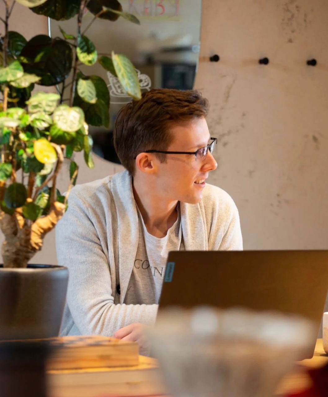 Młody mężczyzna w okularach siedzi przy biurku z laptopem, spoglądając w bok, obok doniczka z rośliną