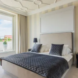 Elegancka sypialnia z łóżkiem typu king-size, ciemnoszarą pikowaną narzutą i widokiem na balkon z zielenią.