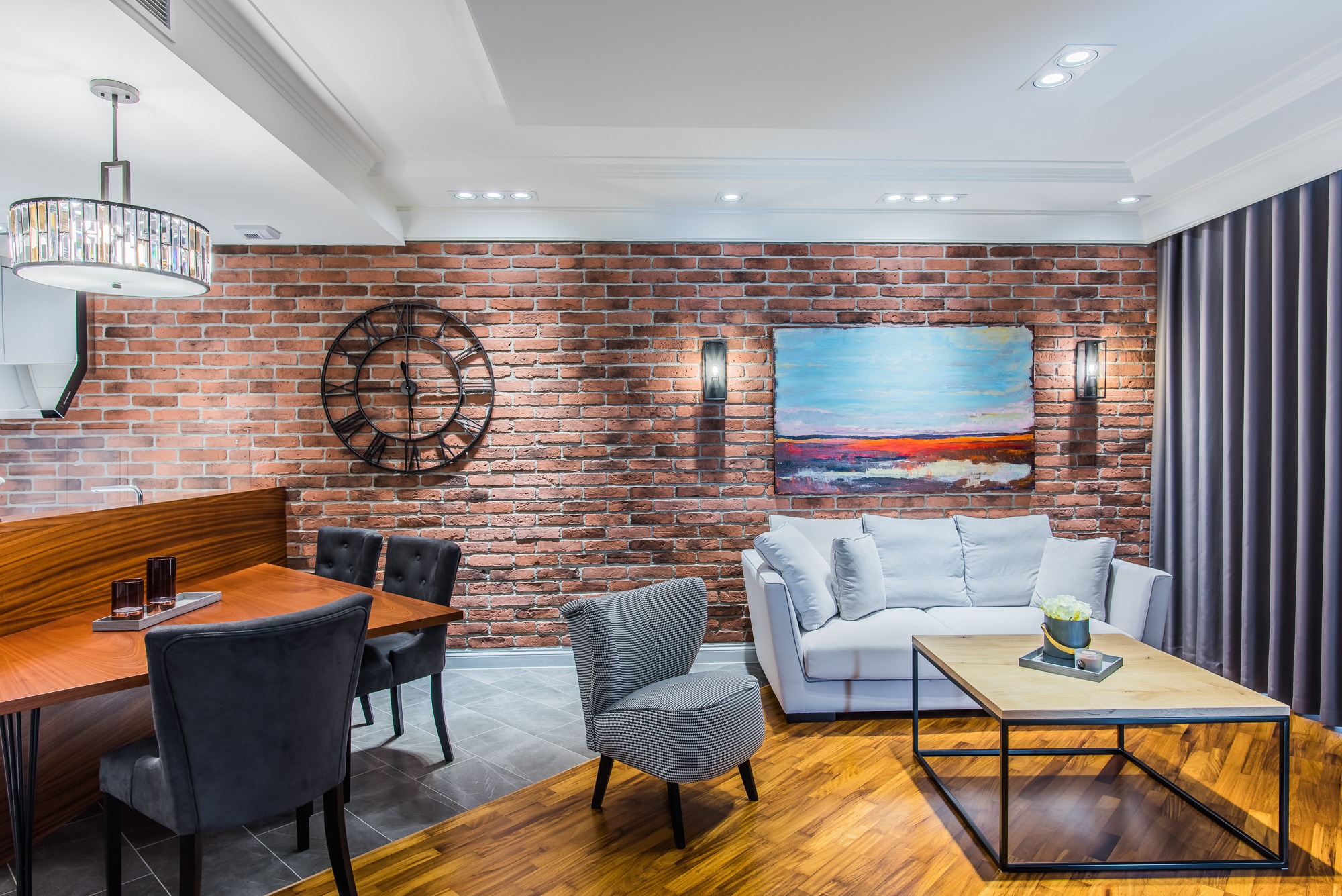 Salon z ceglaną ścianą, szarą sofą, drewnianym stolikiem kawowym i krzesłem w pepitkę, oświetlony lampami ściennymi i wiszącymi.