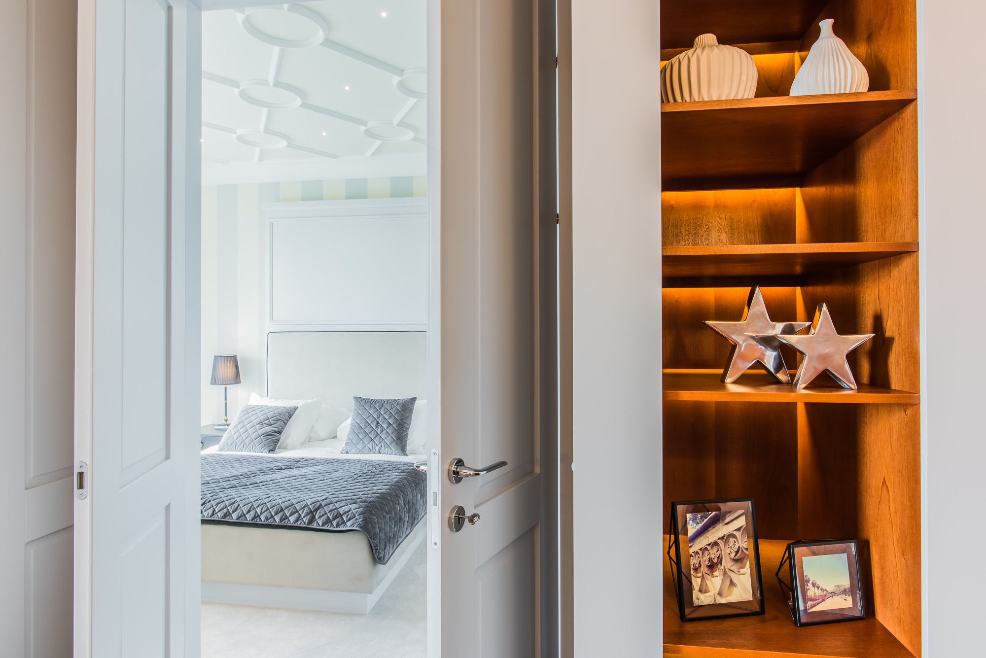 Widok na sypialnię przez uchylone białe drzwi, z drewnianym regałem po prawej stronie, na którym stoją srebrne gwiazdy, ceramiczne wazony i ramki ze zdjęciami.