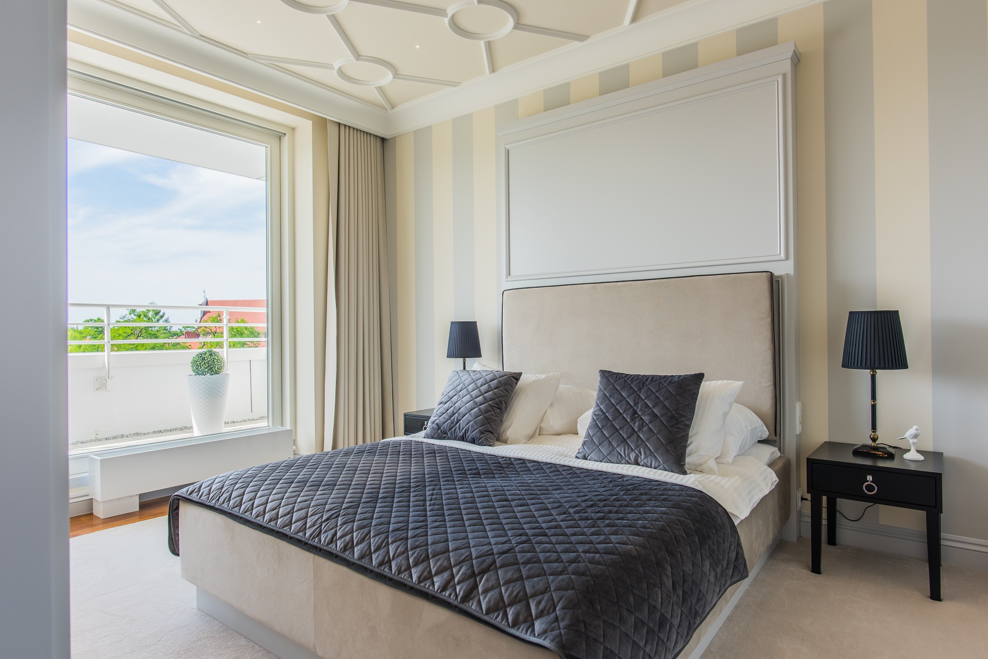 Elegancka sypialnia z łóżkiem typu king-size, ciemnoszarą pikowaną narzutą i widokiem na balkon z zielenią.