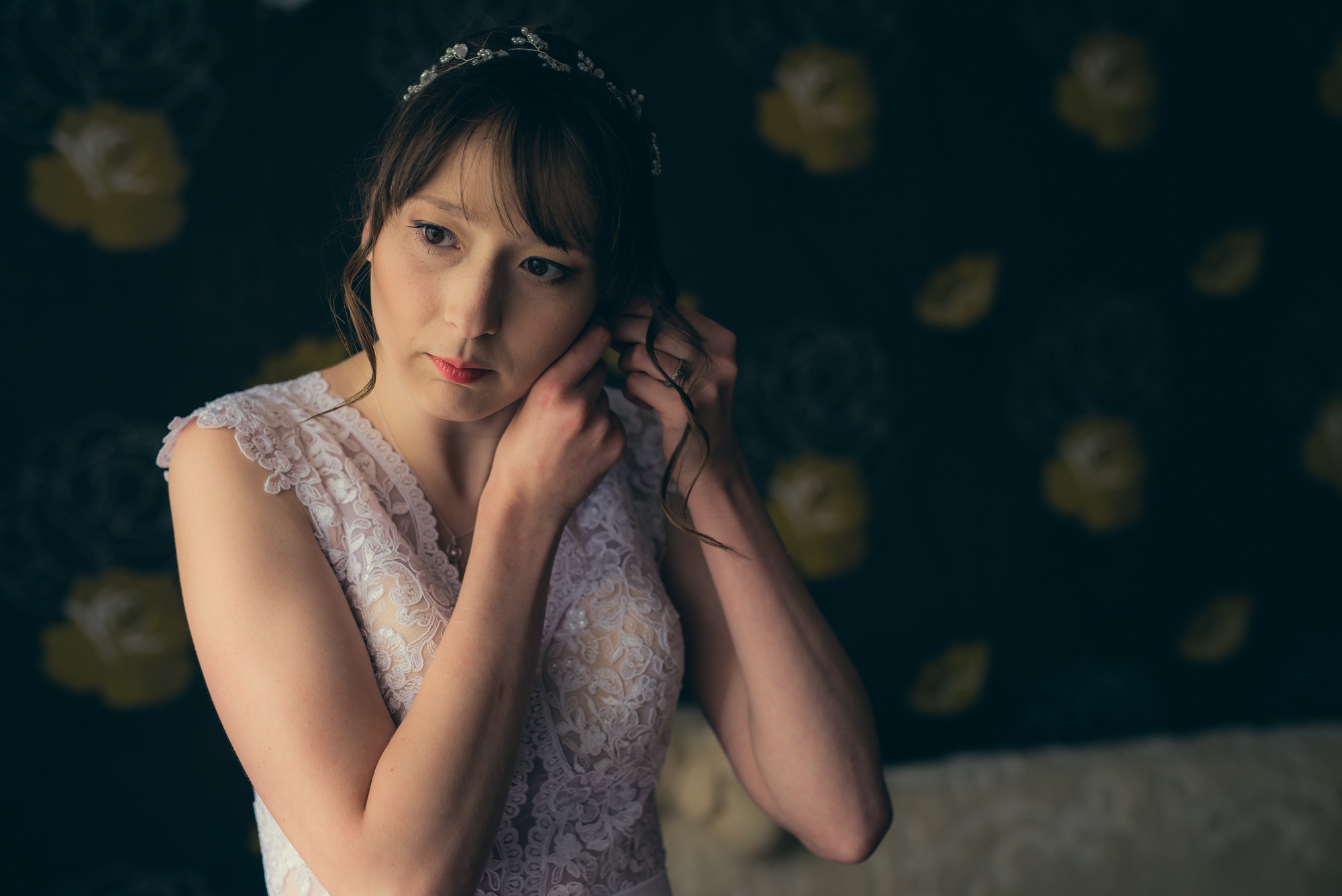 Panna młoda w koronkowej sukni, z diademem we włosach, delikatnie wkłada kolczyk, na ciemnym tle z motywem kwiatowym.