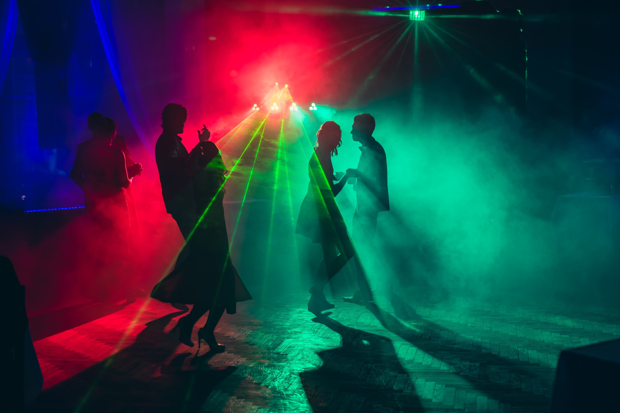 Sylwetki par tańczących w zadymionym pomieszczeniu oświetlonym czerwonymi i zielonymi laserami, parkiet z ciemnego drewna.