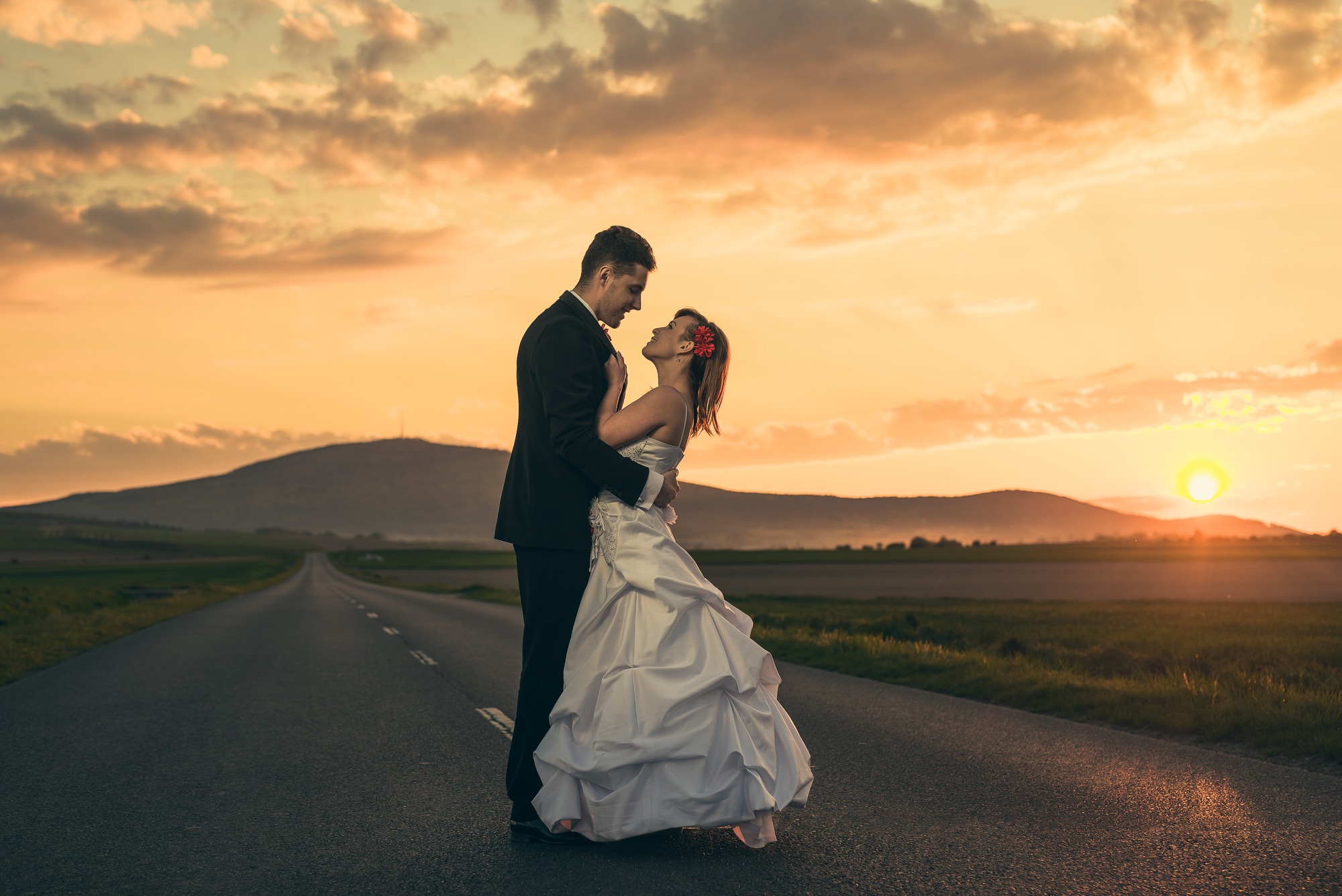 Para młoda obejmująca się na asfalcie drogi w promieniach zachodzącego słońca, widok na horyzont i wzgórza.