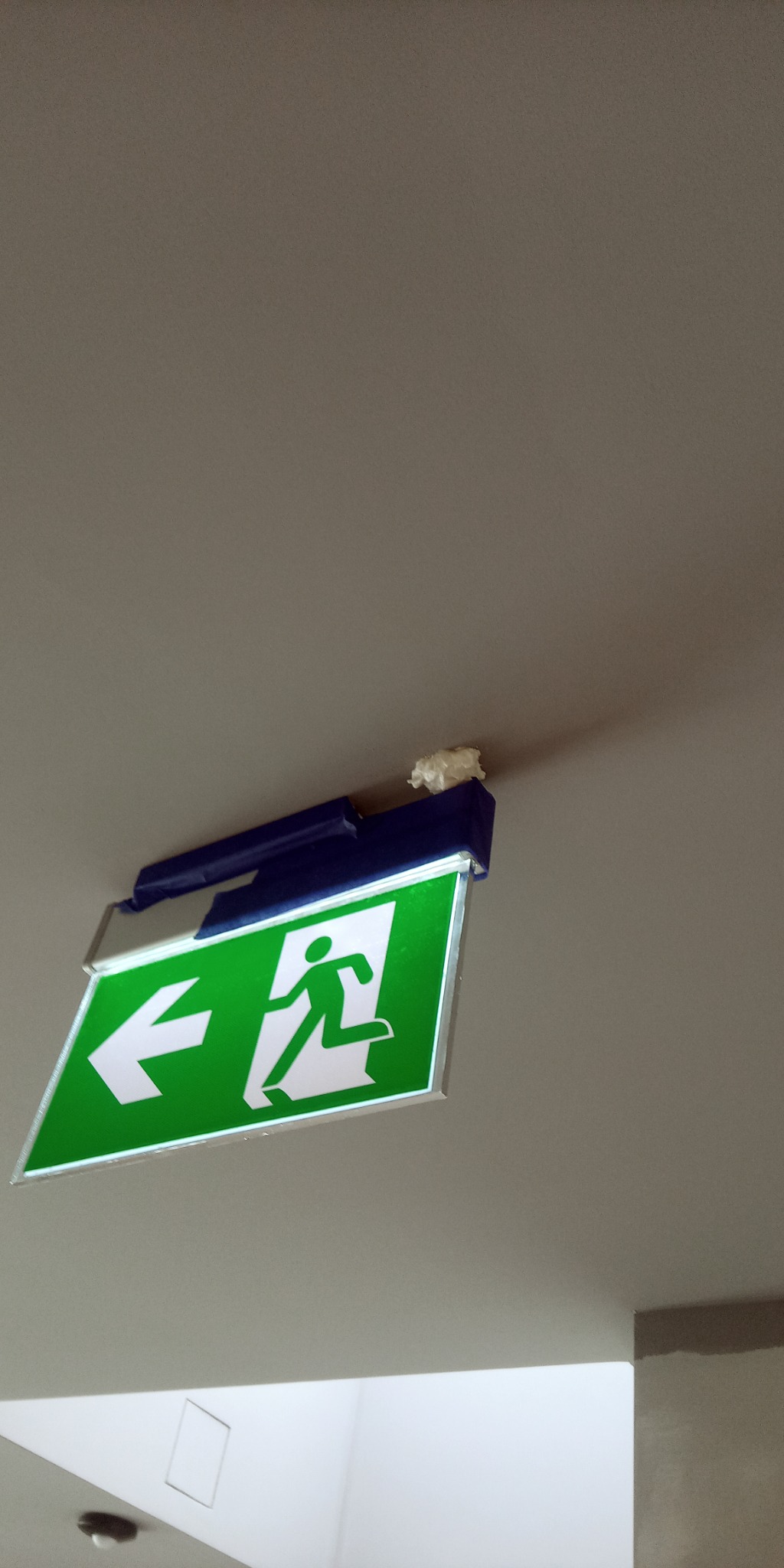 Zielony znak ewakuacyjny z białą strzałką i sylwetką biegnącego człowieka, zamontowany pod sufitem, z widoczną niezabezpieczoną watą izolacyjną.