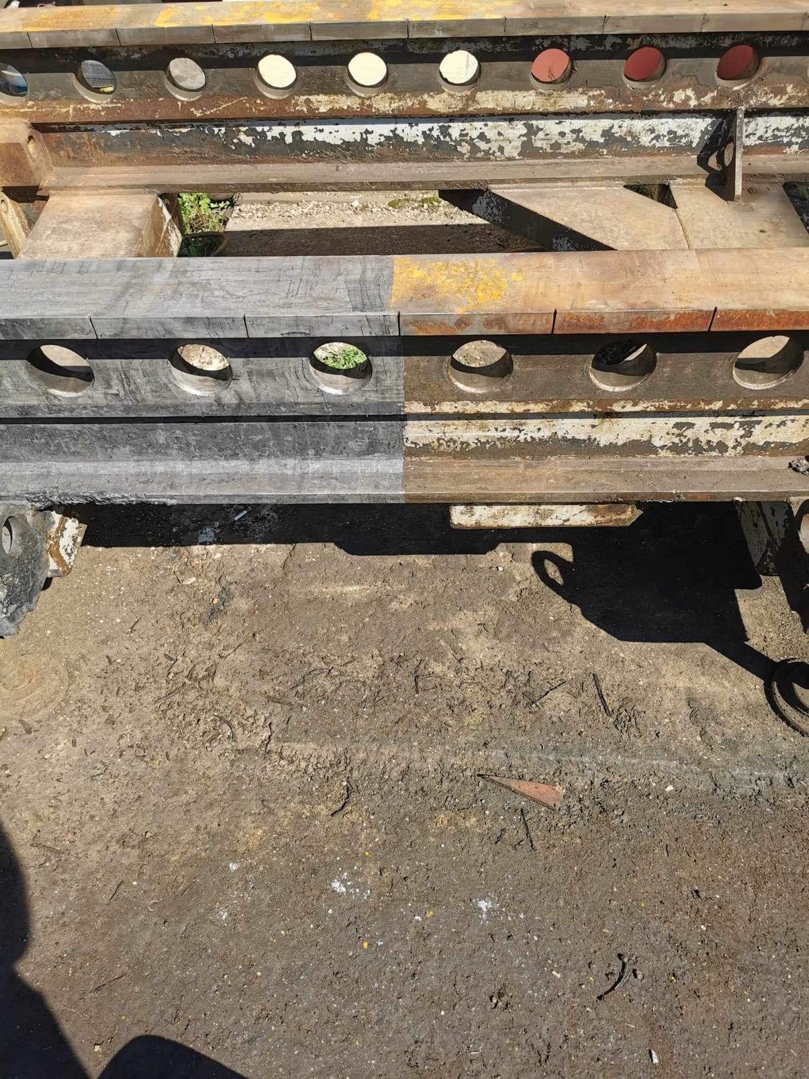 Metalowa konstrukcja z otworami, połowa wyczyszczona, druga pokryta rdzą i starą farbą, leżąca na ziemi.