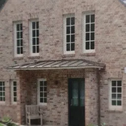 Dwupiętrowy dom z cegły z białymi oknami PCV, półokrągłym oknem na poddaszu i zadaszonym wejściem z ławką.