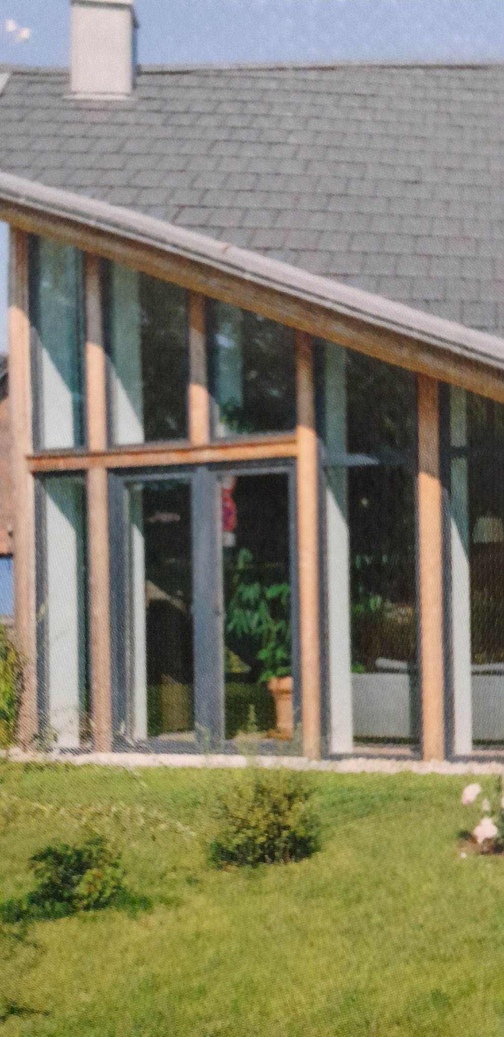 Nowoczesna fasada domu z dużymi oknami i przeszklonymi drzwiami wejściowymi, widok z zewnątrz na trawnik i roślinność.