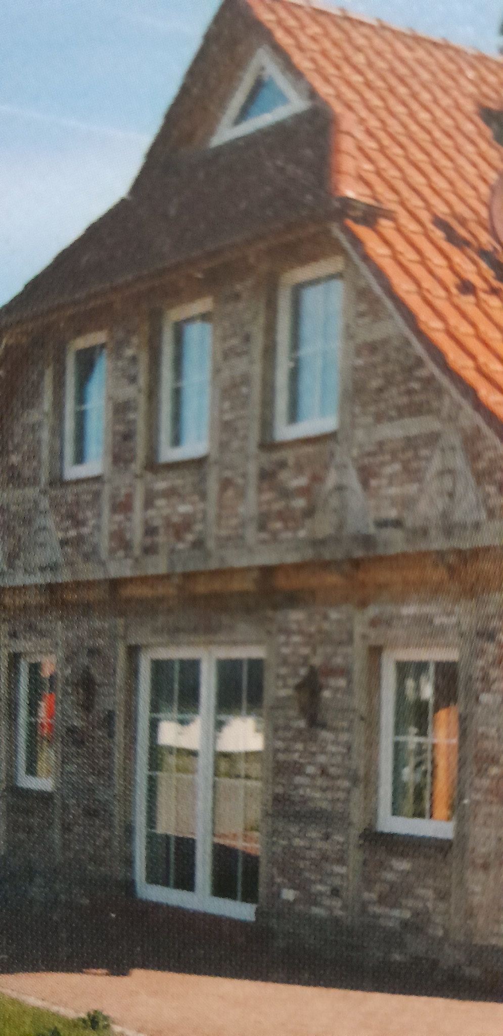 Dwukondygnacyjny dom z cegły klinkierowej z białymi oknami i drzwiami balkonowymi, dachówka w kolorze ceglastym, małe trójkątne okno na poddaszu.