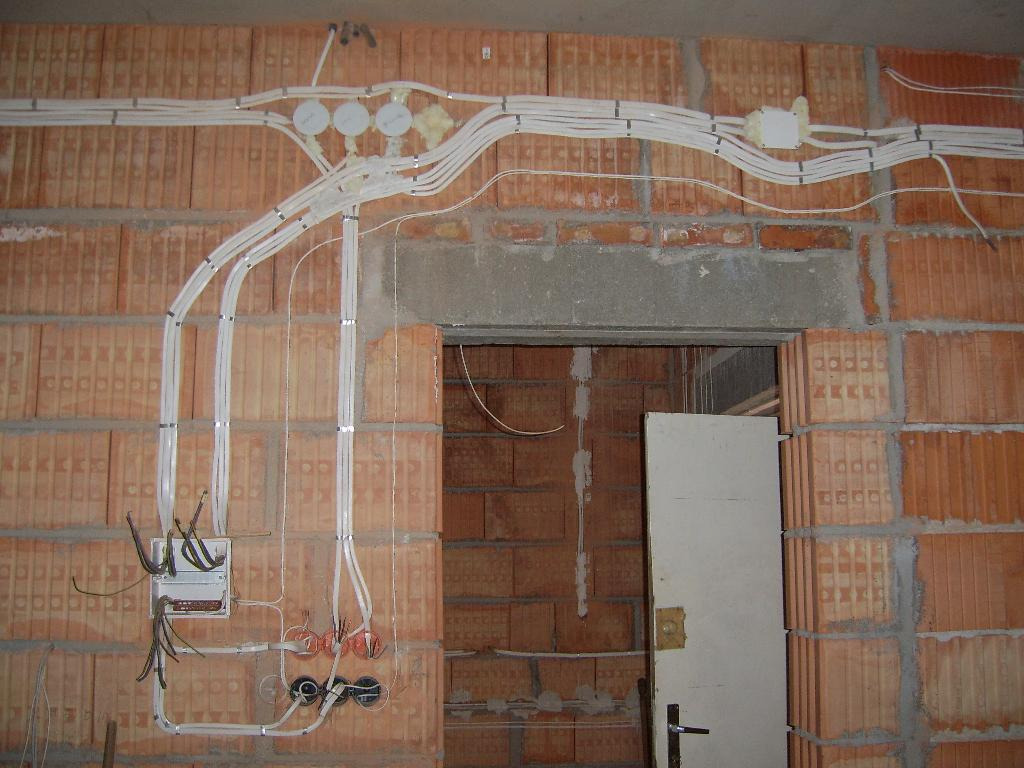 Surowa ceglana ściana z widocznymi, nowo położonymi białymi przewodami elektrycznymi, rozdzielającymi się do puszek instalacyjnych; widoczne kable wychodzące ze skrzynki elektrycznej i przejście...