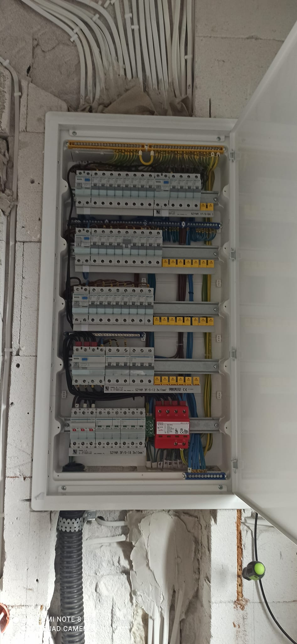 Otwarta skrzynka elektryczna z zamontowanymi bezpiecznikami, przewodami i listwami zaciskowymi na surowej ścianie z widocznymi przewodami w peszlach.