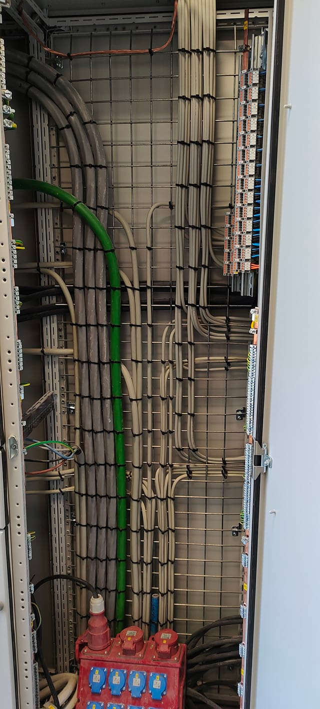 Rozdzielnica elektryczna z uporządkowanymi kablami w peszlach i listwach, widoczne gniazda siłowe na dole obudowy, metalowa siatka wewnątrz obudowy.