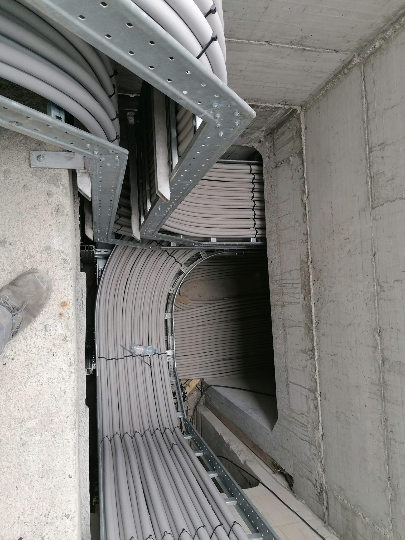 Rzut z góry na koryto kablowe w surowym betonowym szybie, wypełnione równolegle ułożonymi szarymi kablami, zabezpieczonymi czarnymi opaskami zaciskowymi; w tle ciemny otwór, a w lewym dolnym rogu...