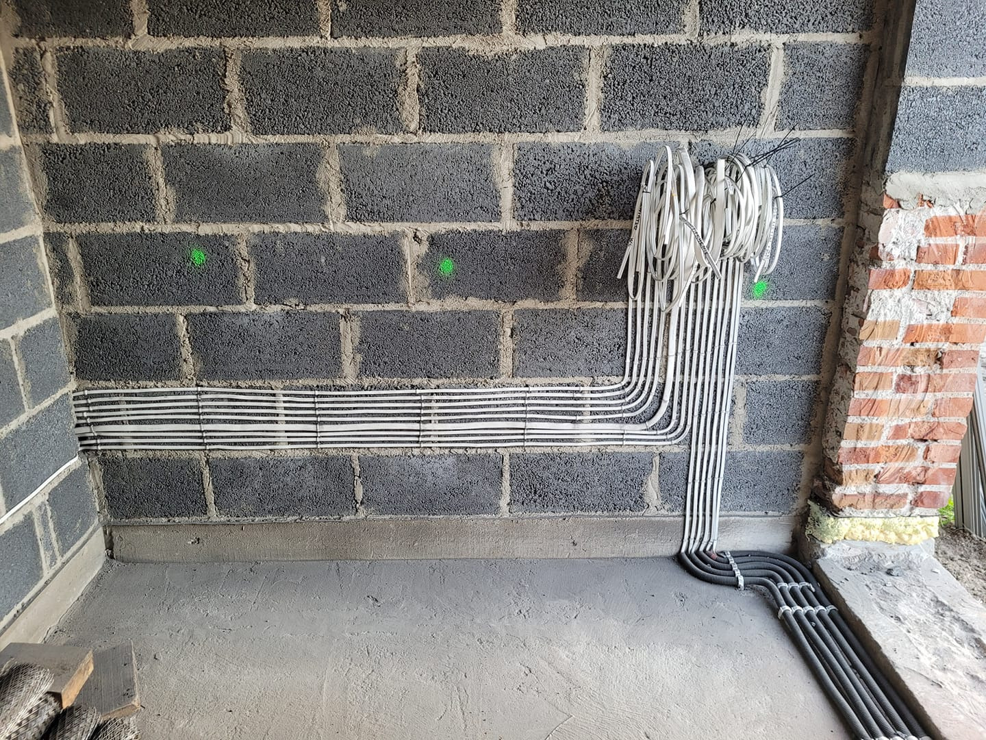 Surowe wnętrze z szarego betonu komórkowego. Widoczne ułożone poziomo i pionowo kable elektryczne w peszlach oraz wiązka białych przewodów przymocowana do ściany. Dwa zielone punkty na ścianie.