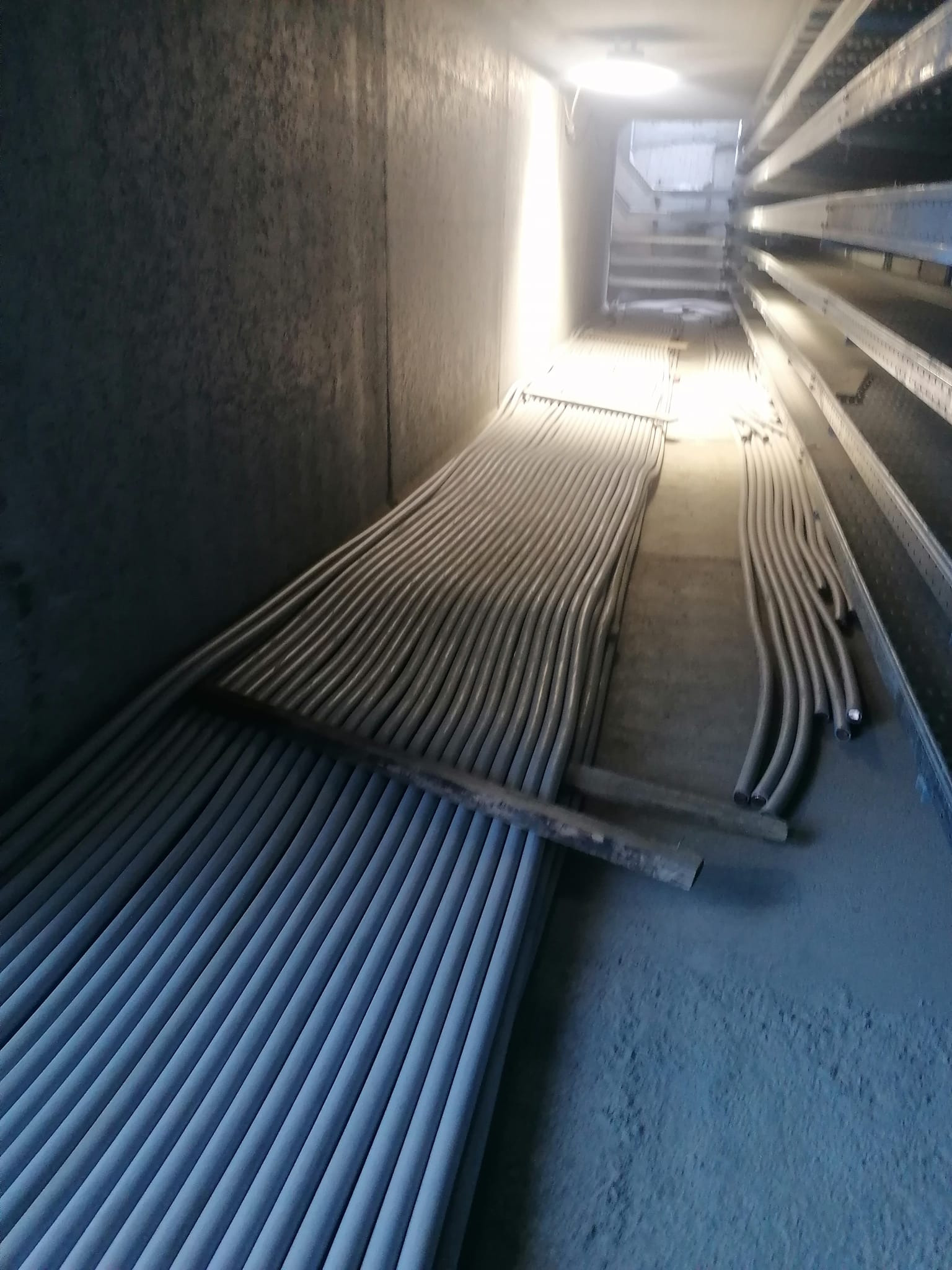 Rzędy ułożonych kabli elektrycznych w szarym, betonowym korytarzu z metalowymi półkami po prawej stronie, oświetlonym górnym światłem.