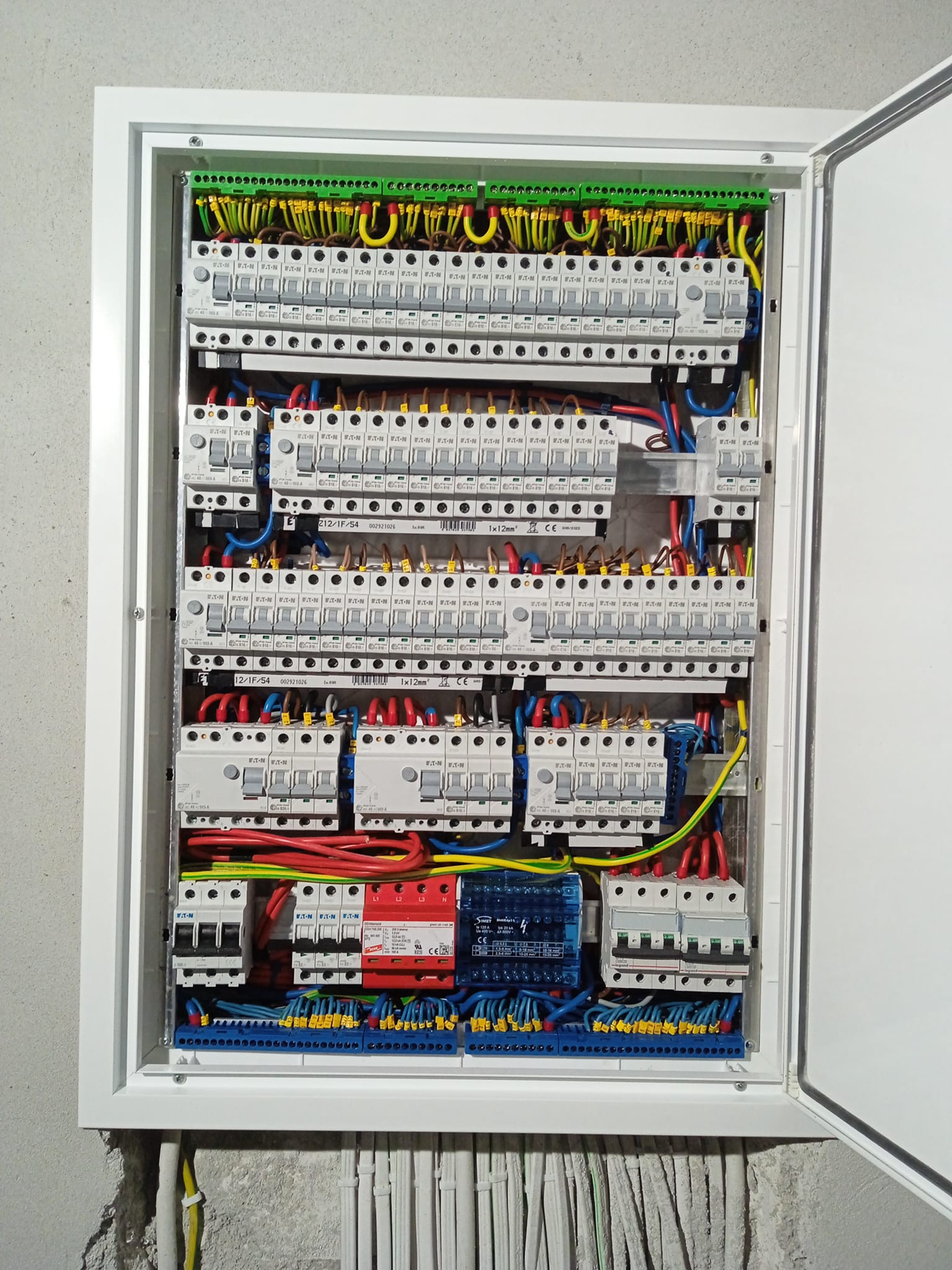 Otwarta skrzynka elektryczna z licznymi bezpiecznikami, przewodami w różnych kolorach (żółty, zielony, niebieski, czerwony) i listwami zaciskowymi, zamontowana na szarej, surowej ścianie.