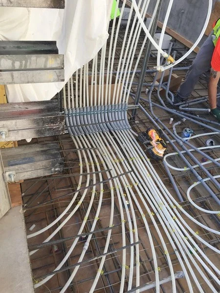Rozprowadzenie białych rurek instalacji ogrzewania podłogowego w siatce zbrojeniowej, widoczne złącza i narzędzie do montażu, fragment posadzki w trakcie prac.