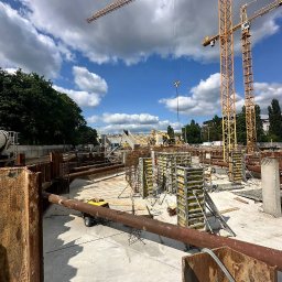 BELBUDPRO Sp. z o.o. - Budowa fundamentów w Poznaniu: widok na plac budowy z widocznymi elementami zbrojenia, szalunkami i dźwigami. Prace ziemne w toku, widoczne rury i maszyny.