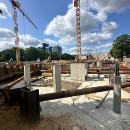 BELBUDPRO Sp. z o.o. - Betonowe filary na placu budowy z żurawiami w tle, widok w słoneczny dzień. Metalowe rury i zbrojenia na pierwszym planie.