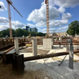 Betonowe filary na placu budowy z żurawiami w tle, widok w słoneczny dzień. Metalowe rury i zbrojenia na pierwszym planie.