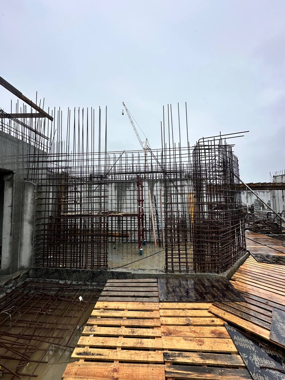 Zbrojenie pionowe i poziome na budowie, widoczne drewniane palety i żuraw budowlany w tle, pochmurne niebo.