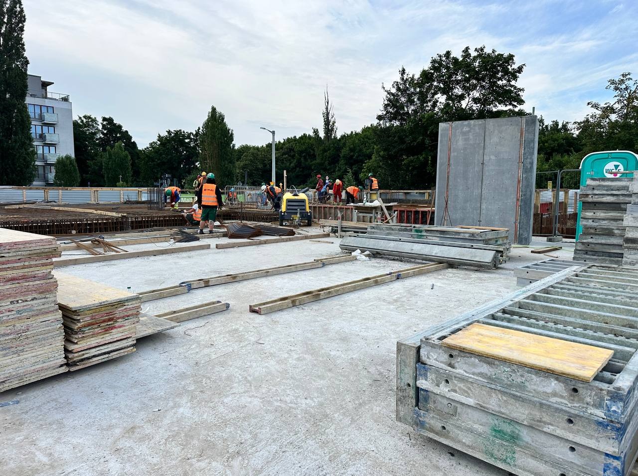 Budowa w toku: pracownicy w odblaskowych kamizelkach montują zbrojenie na betonowej płycie fundamentowej, widoczne szalunki i sprzęt budowlany.