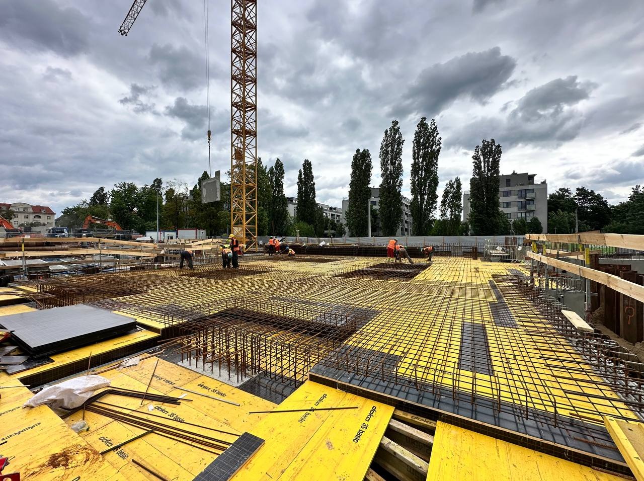Żelbetowa konstrukcja w budowie, widoczne zbrojenia na żółtym szalunku, pracownicy w pomarańczowych kamizelkach i dźwig na tle pochmurnego nieba.