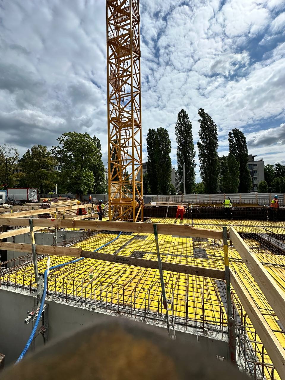 Żelbetowa płyta fundamentowa z żółtym szalunkiem i zbrojeniem, przygotowana do zalania betonem. Na budowie widoczny żuraw wieżowy i pracownicy w kaskach.