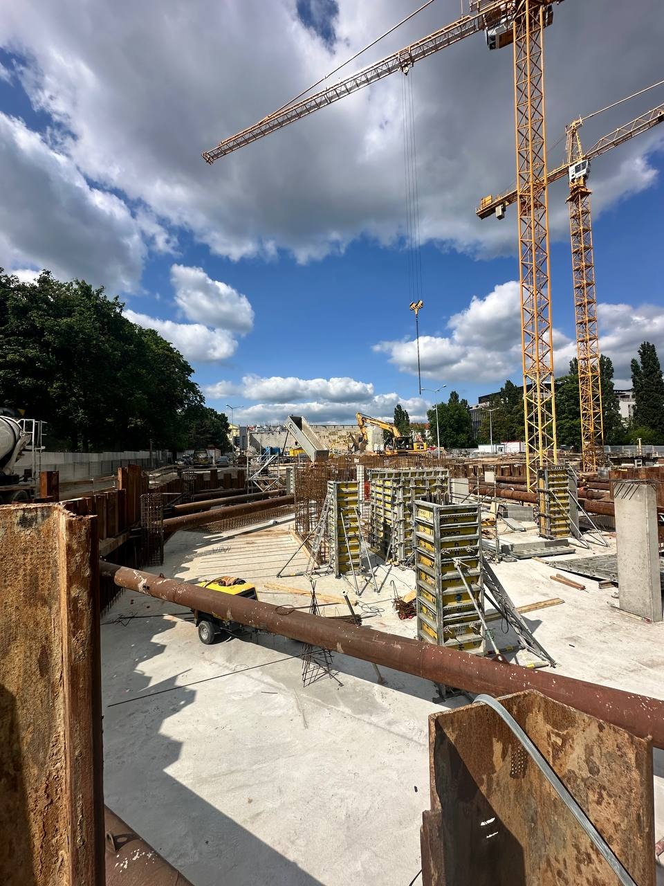 Budowa fundamentów w Poznaniu: widok na plac budowy z widocznymi elementami zbrojenia, szalunkami i dźwigami. Prace ziemne w toku, widoczne rury i maszyny.