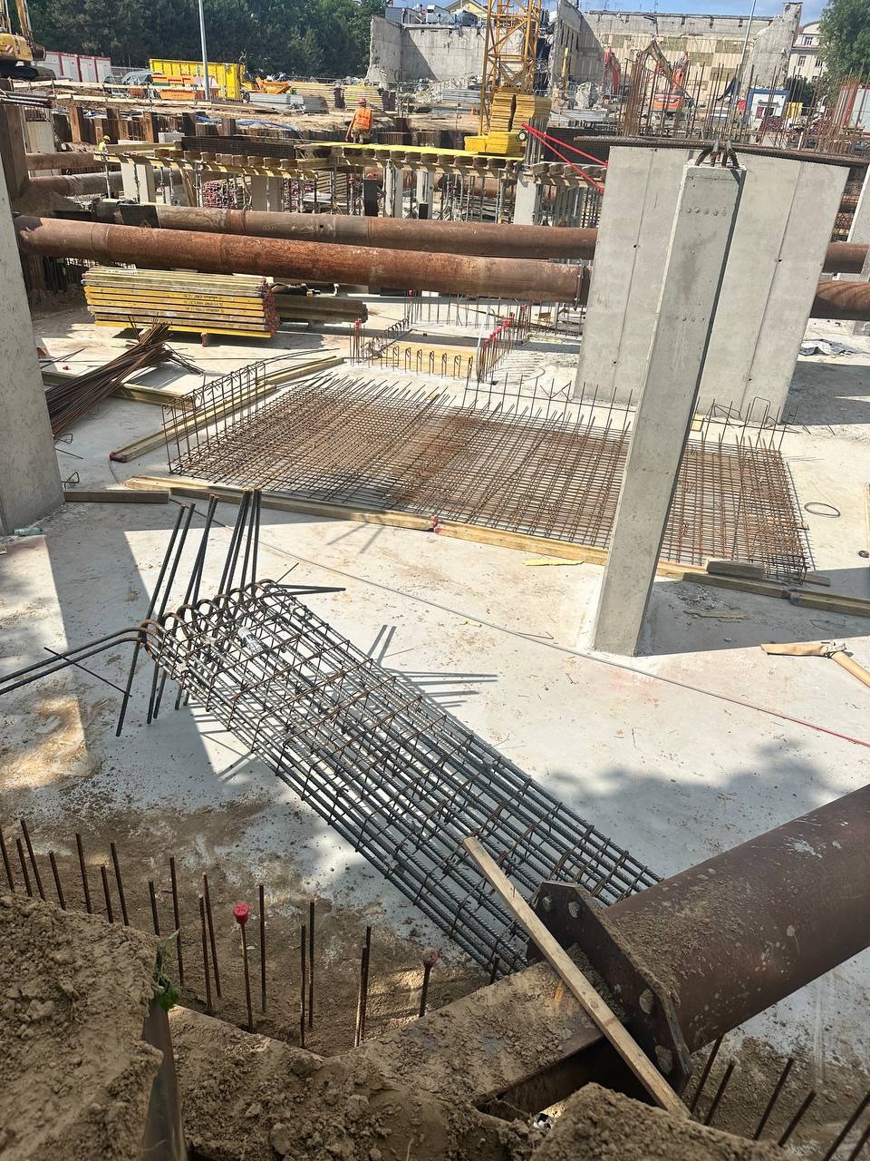 Zbrojenie fundamentów na budowie: widok z góry na siatkę zbrojeniową, stalowe słupy i rury na placu budowy w słoneczny dzień. W tle widoczny dźwig.