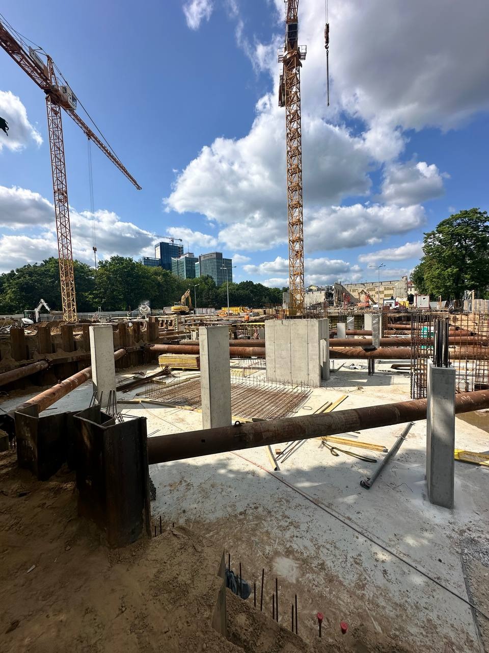 Betonowe filary na placu budowy z żurawiami w tle, widok w słoneczny dzień. Metalowe rury i zbrojenia na pierwszym planie.