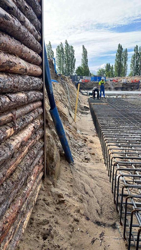 Budowa fundamentów: drewniana ścianka oporowa, zbrojenie stalowe i pracownicy w oddali przygotowujący beton.