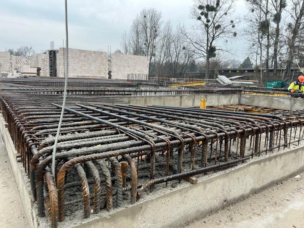 Zbrojenie fundamentu domu w trakcie budowy, widoczne stalowe pręty związane drutem, szalunek betonowy i pracownik w oddali.