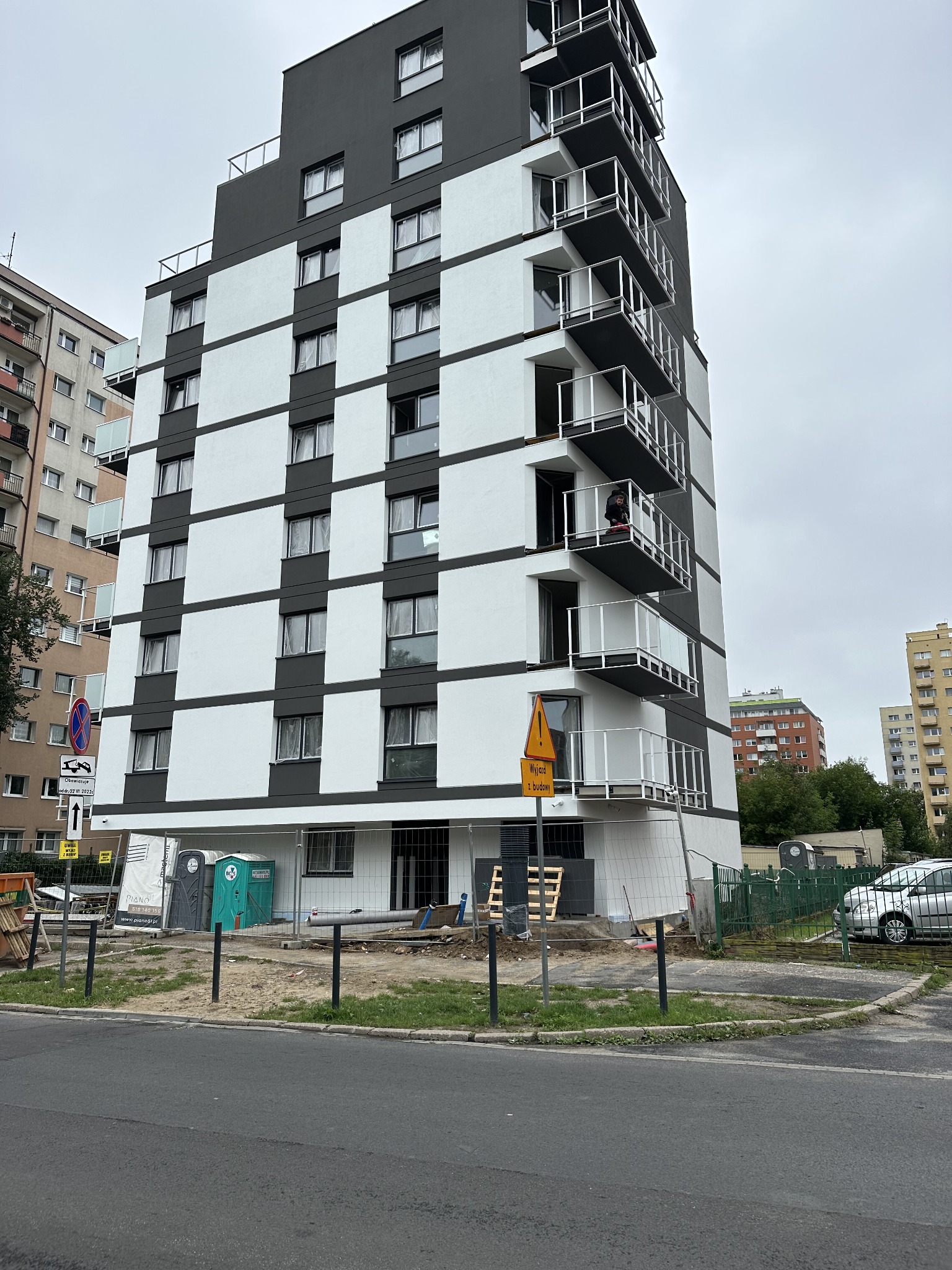 Wielopiętrowy budynek mieszkalny w trakcie budowy, z widocznymi balkonami z białymi balustradami, szara elewacja z białymi panelami, teren budowy z tymczasowymi toaletami i ogrodzeniem, widoczny...