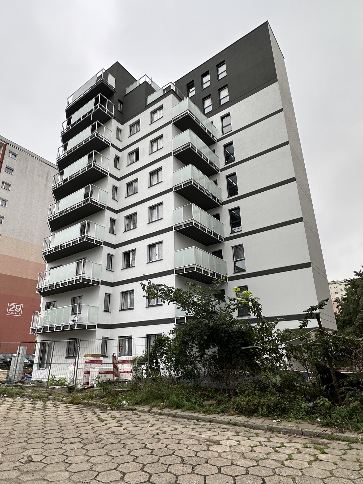 Wielopiętrowy budynek mieszkalny z balkonami z białymi balustradami szklanymi, w trakcie wykończenia terenu wokół budynku, widoczne tymczasowe ogrodzenie i materiały budowlane.