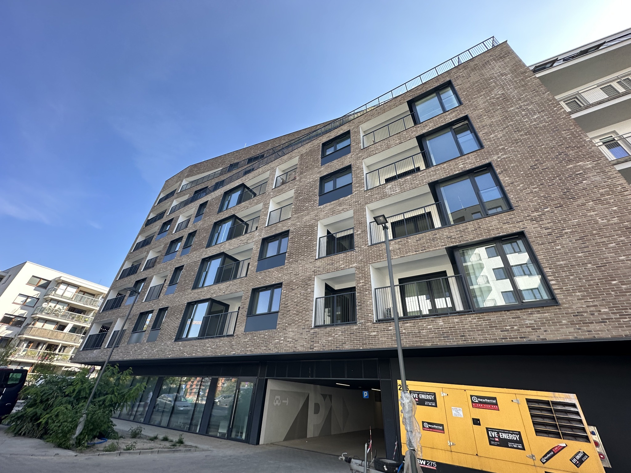 Nowoczesny budynek mieszkalny z balkonami i podziemnym parkingiem, widoczny agregat prądotwórczy na pierwszym planie.