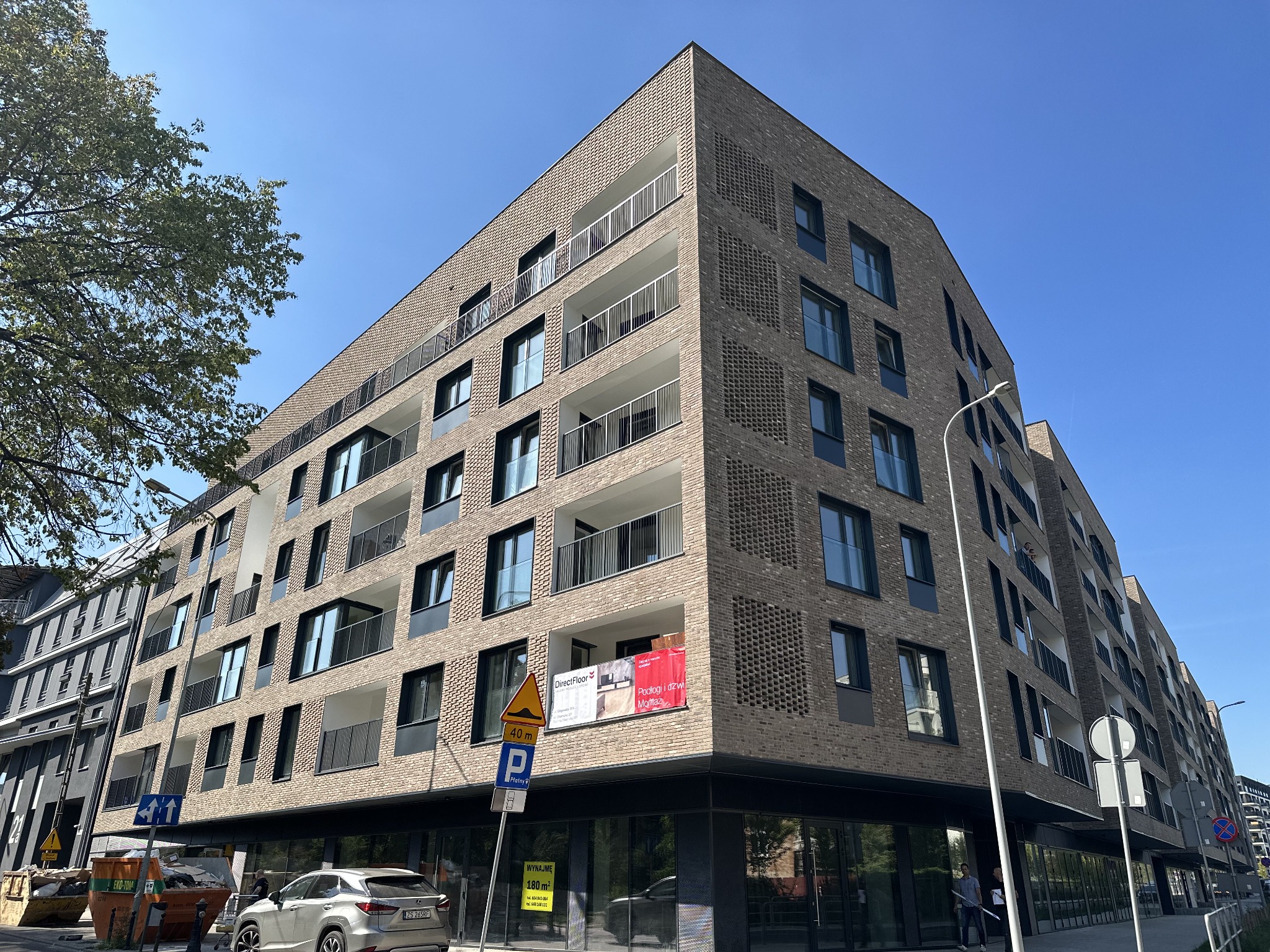 Nowoczesny budynek mieszkalny w Poznaniu z balkonami zabezpieczonymi metalowymi balustradami, wykończony cegłą w odcieniach szarości i brązu, z widocznymi szyldami reklamowymi na elewacji.