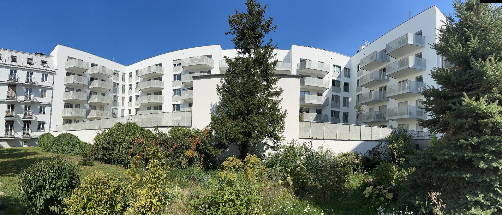 Nowoczesny budynek apartamentowy z białymi balkonami i przeszklonymi balustradami, otoczony zielenią w słoneczny dzień.