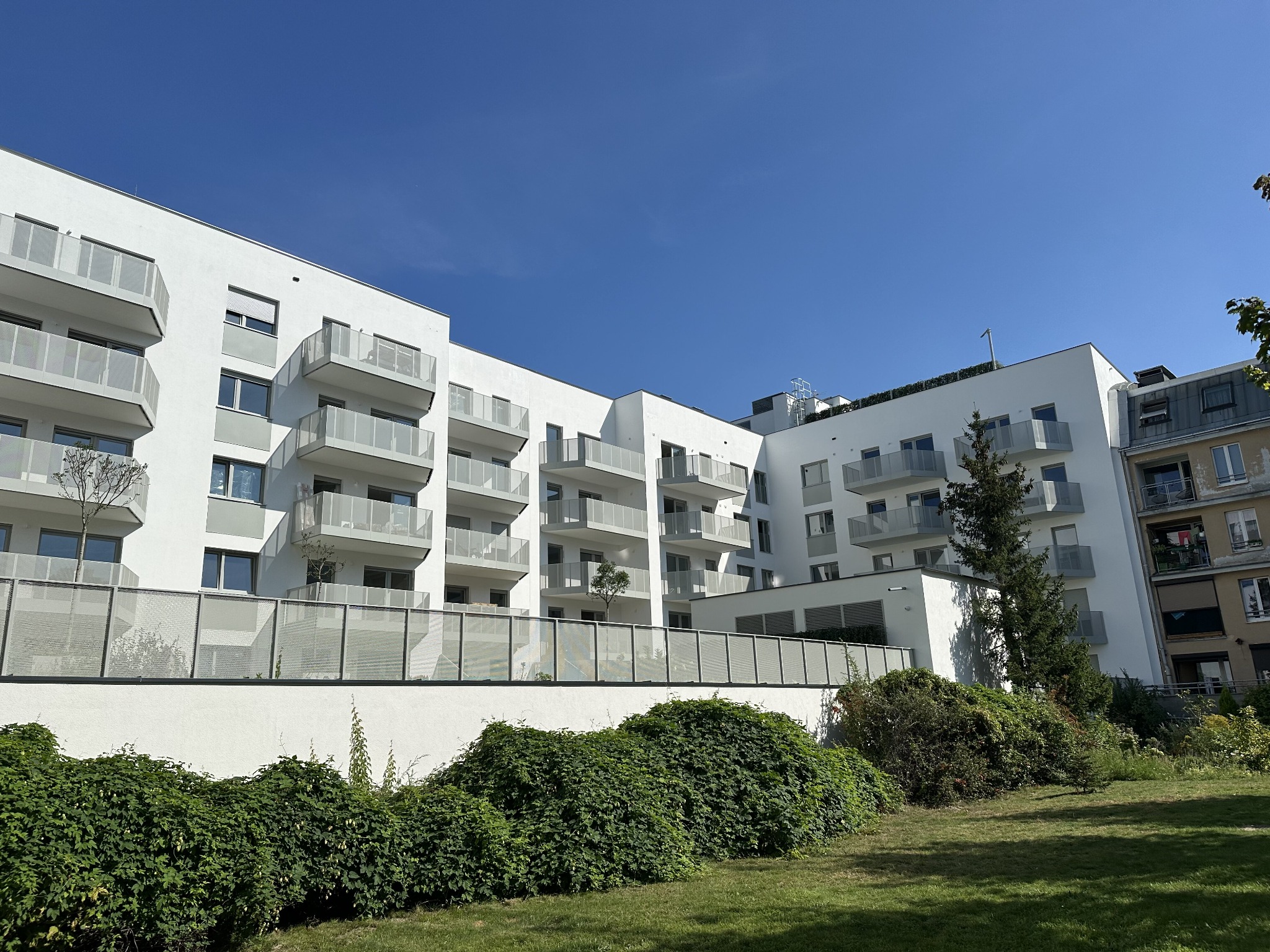 Nowoczesny, biały budynek mieszkalny z balkonami o szklanych balustradach, widok z dołu na tle błękitnego nieba, zieleń w dolnej części kadru.