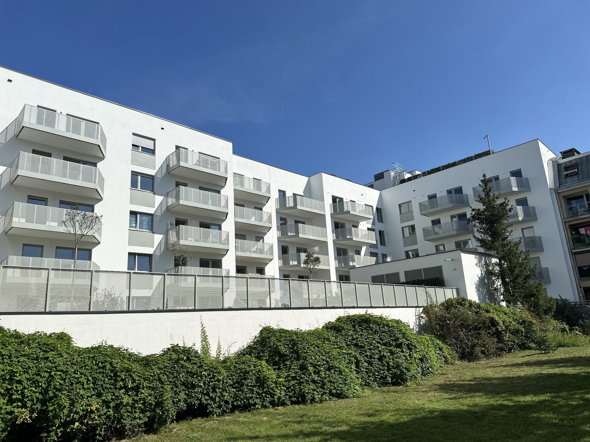 Nowoczesny, biały budynek wielorodzinny z balkonami z szklanymi balustradami, widok z dołu na tle błękitnego nieba, zieleń w tle.