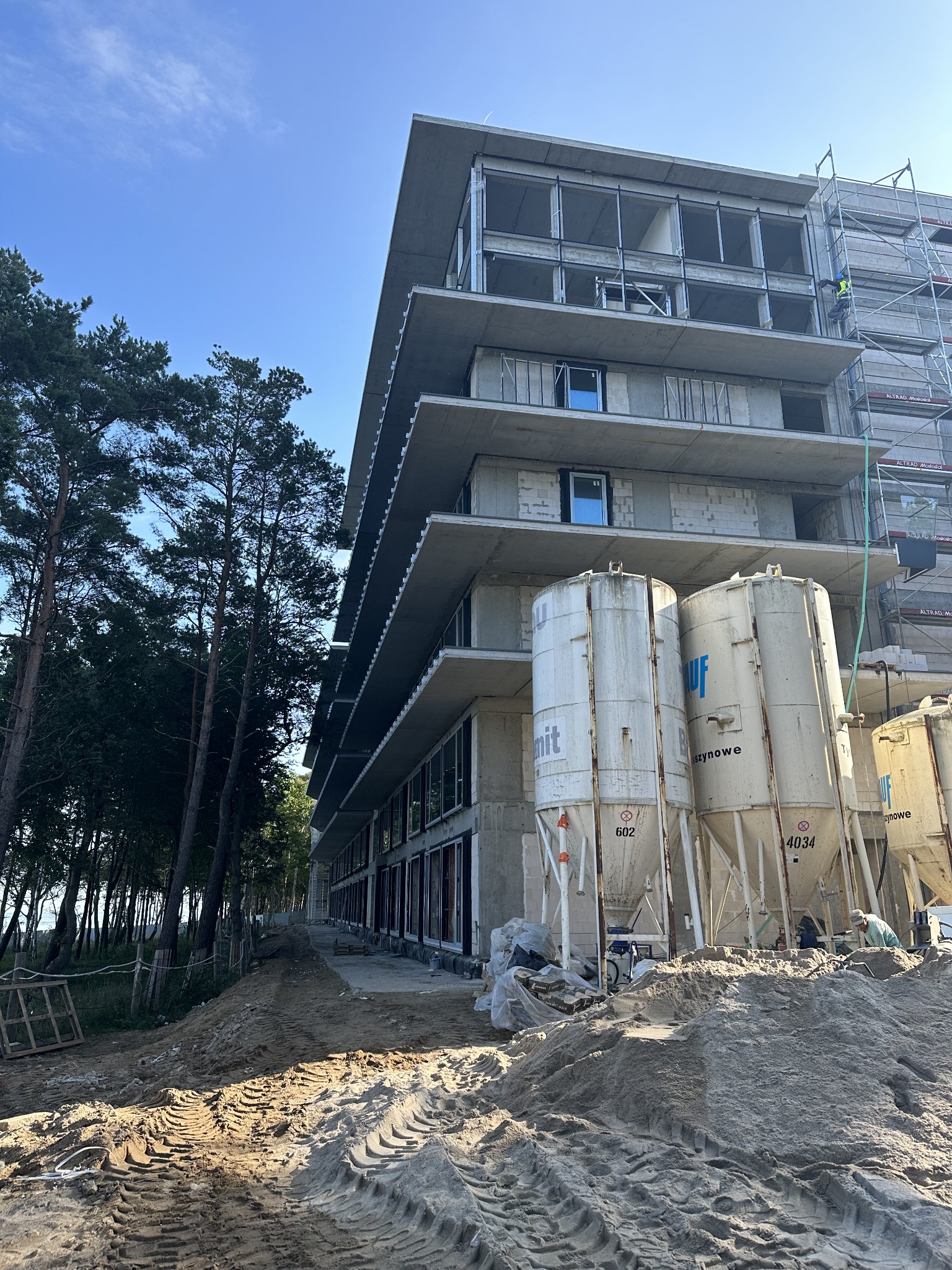 Wznoszący się, surowy budynek w stanie deweloperskim z widocznymi silosami na cement i śladami opon na piaszczystym terenie budowy, zalesione tło i częściowo zamontowanymi oknami.