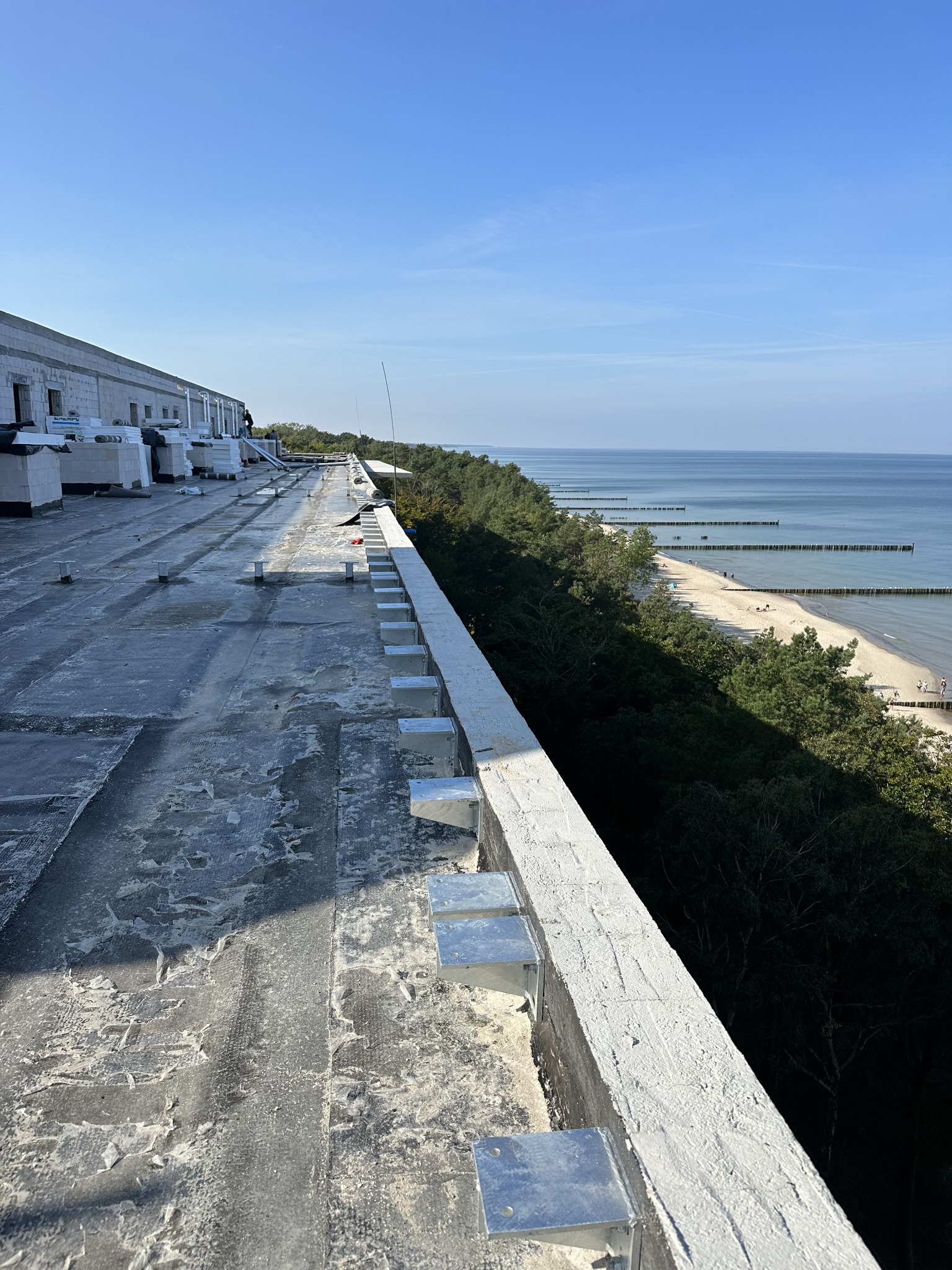 Widok z dachu budynku w trakcie budowy, z widocznym morzem i plażą w tle. Na dachu widoczne materiały izolacyjne i metalowe wsporniki zamontowane na krawędzi.