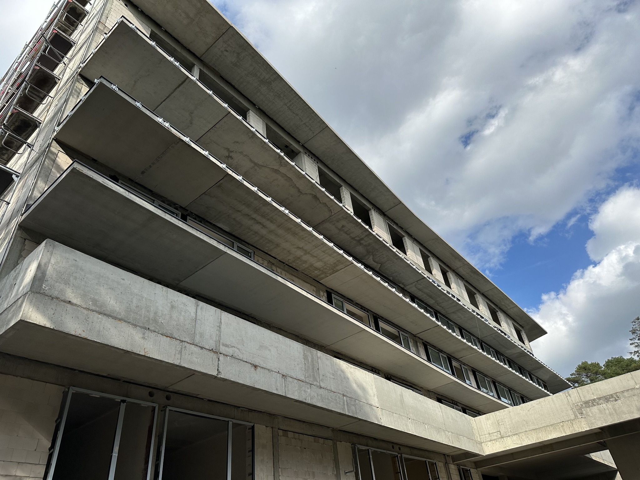 Surowy, betonowy budynek w trakcie budowy, widok z dołu na balkony i okna bez ram, na tle pochmurnego nieba.