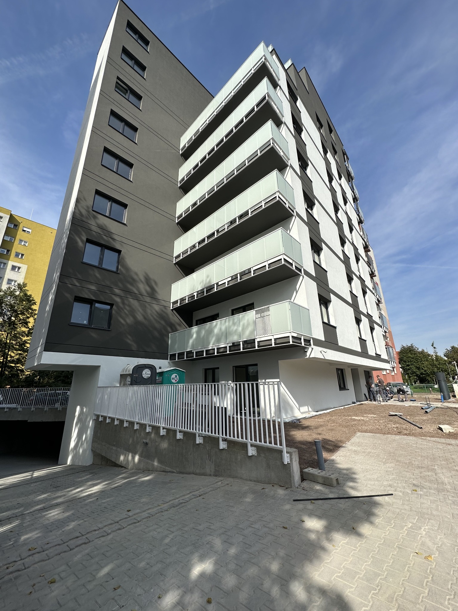 Wielopiętrowy budynek mieszkalny z balkonami zabezpieczonymi szkłem, widok z dołu na elewację w odcieniach szarości i bieli, z widocznymi pracownikami na placu budowy.