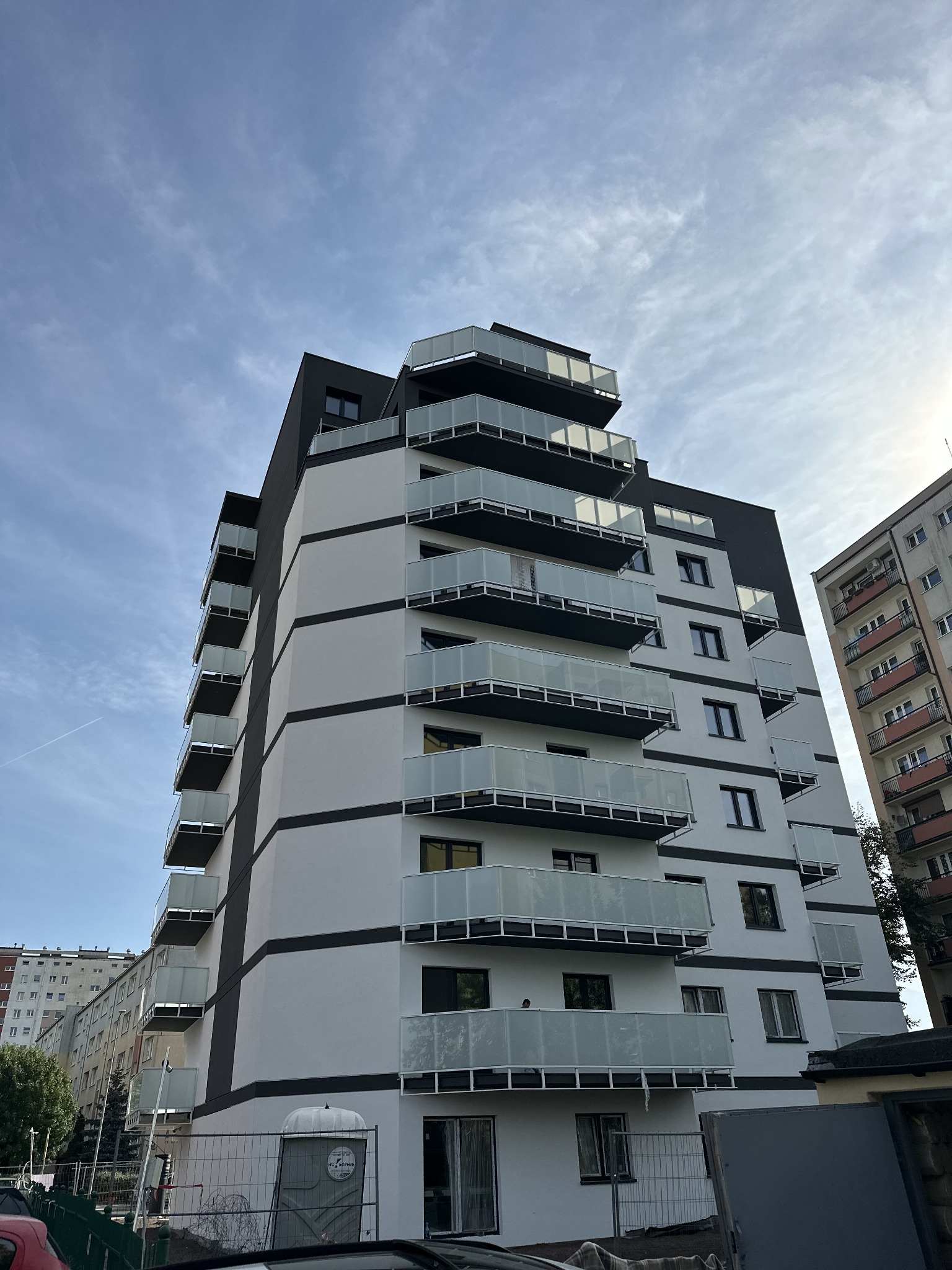 Nowoczesny budynek wielorodzinny z balkonami z balustradami szklanymi, widok z dołu na tle błękitnego nieba z smugą kondensacyjną samolotu.